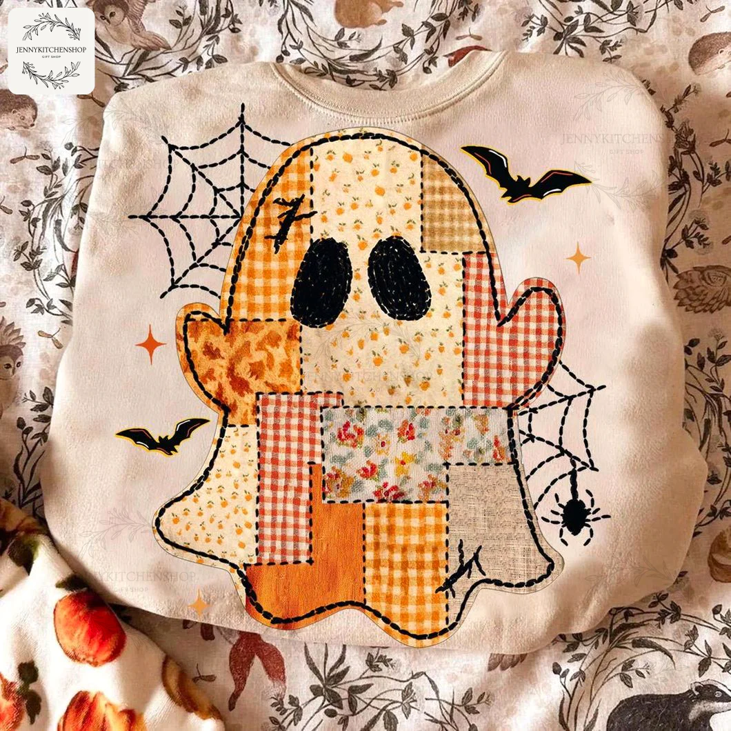 A cream colored sweatshirt with a preppy Halloween patchwork ghost design, laid on a cozy patterned blanket for a fall aesthetic.