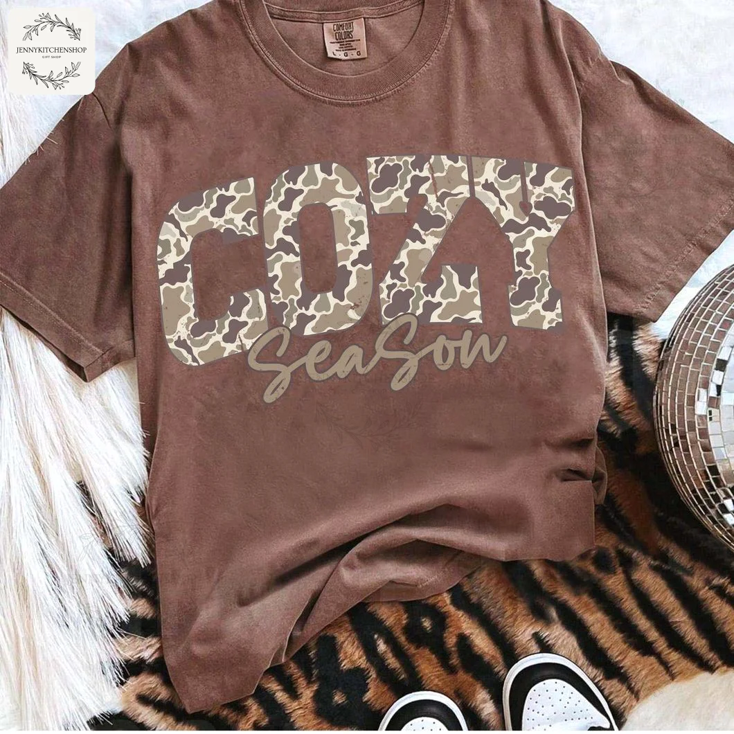 A flat lay of the brown Cozy Season Camo T-Shirt styled on a tiger-print rug with a disco ball and pampas grass.
