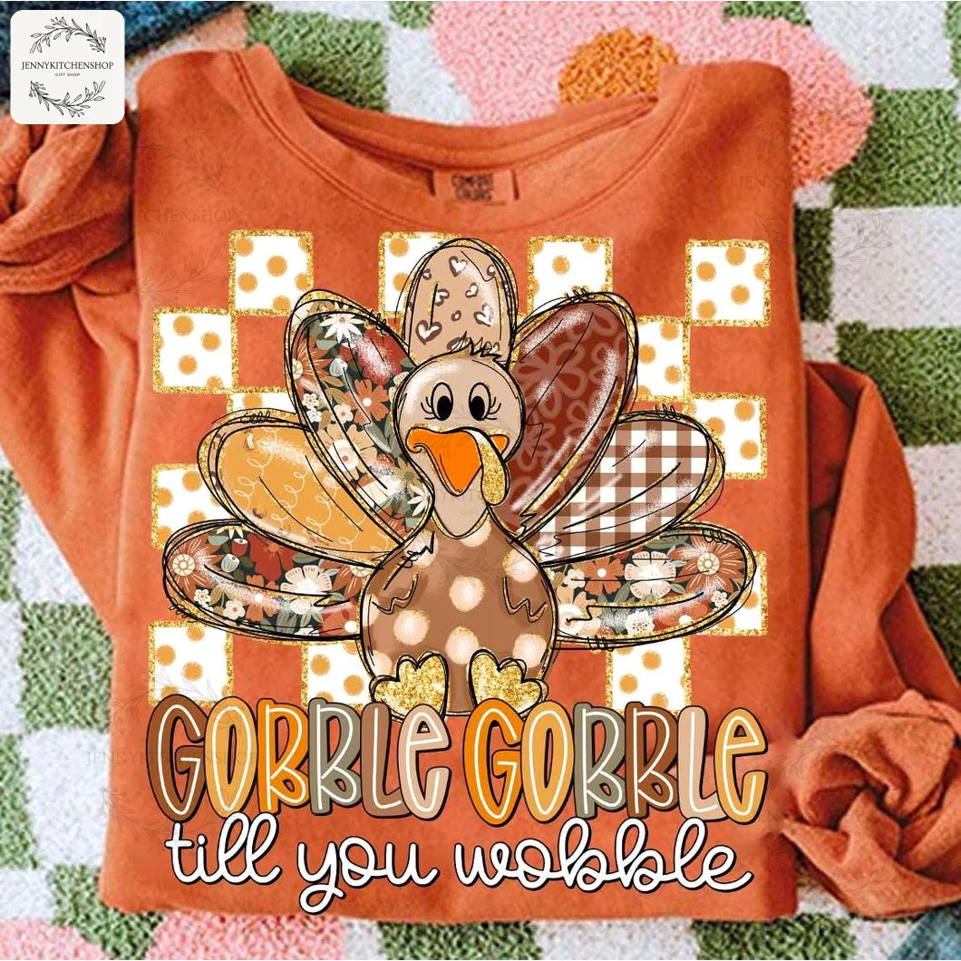 An orange long-sleeve Thanksgiving shirt laid flat, featuring a cute cartoon turkey with patchwork feathers and fun text.